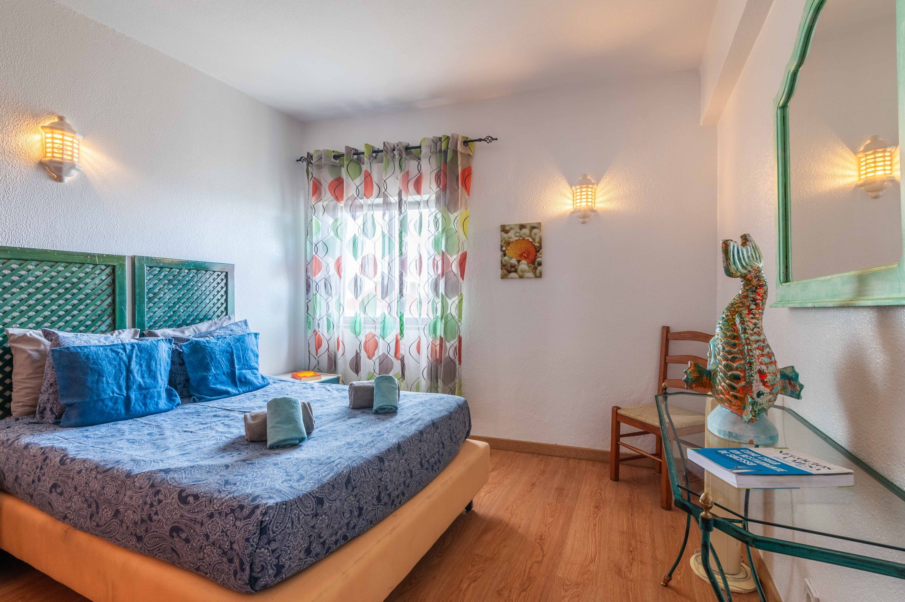 Una camera da letto con un letto, cuscini, una sedia, un tavolino in vetro, una lampada e un oggetto decorativo.