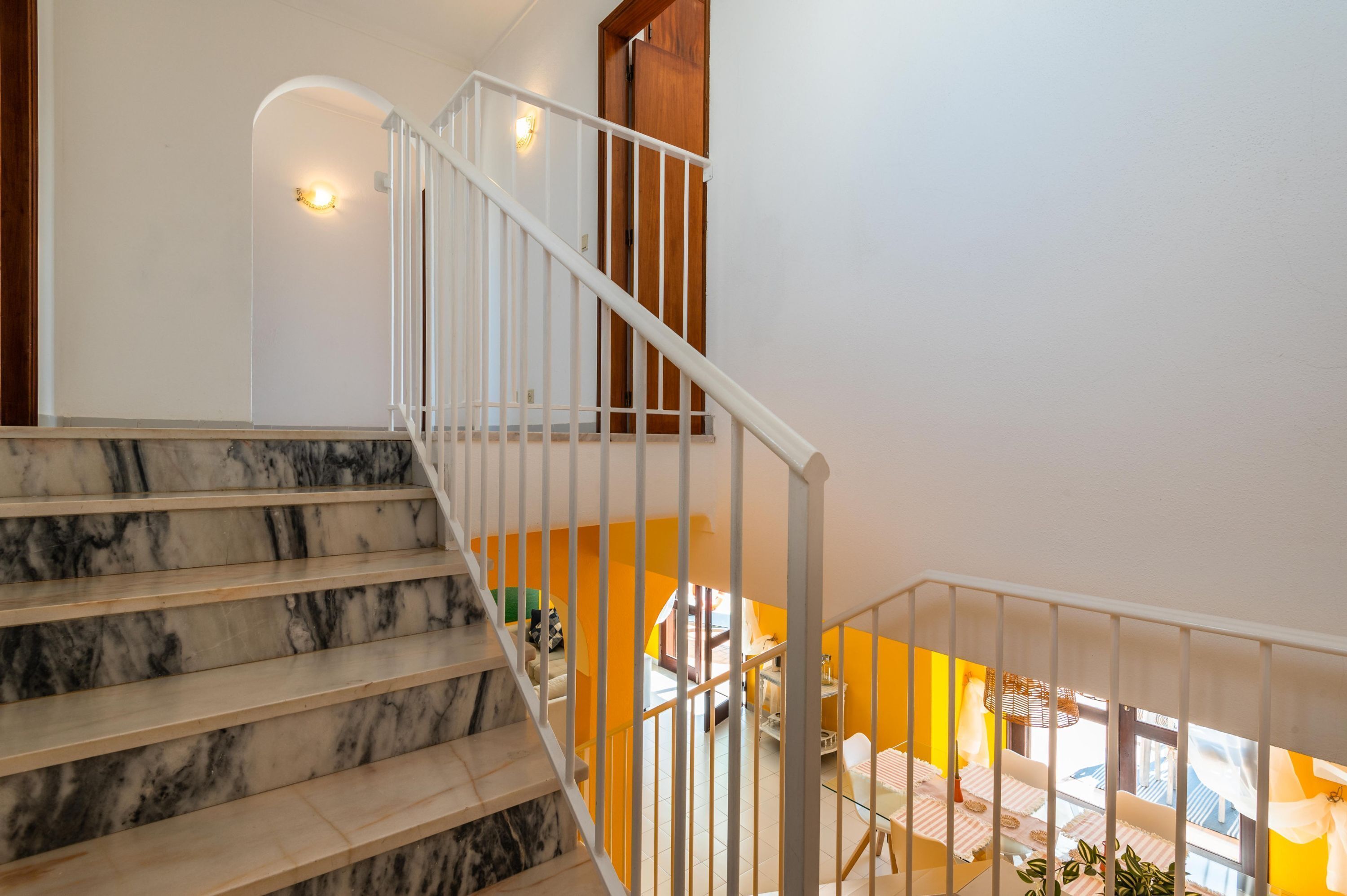 A marble staircase with white railings leading to a second-floor area with yellow walls and a view of the outside.