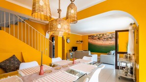 Een helder gele kamer met een eettafel gedekt voor vier personen, witte stoelen, een trap en uitzicht op een strand door een geopende deur.