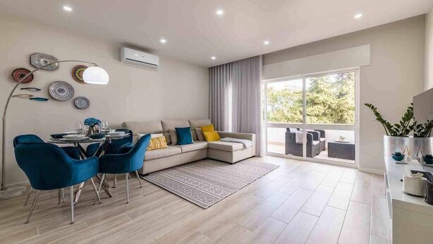Ein modernes Wohnzimmer mit Essbereich, einer Couch mit Kissen, einem Couchtisch und einem großen Fenster mit Blick auf Grünflächen.