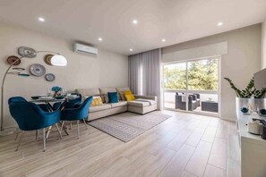 Un salon moderne comprenant un coin repas, un canapé avec des coussins, une table basse et une grande fenêtre donnant sur un paysage verdoyant.