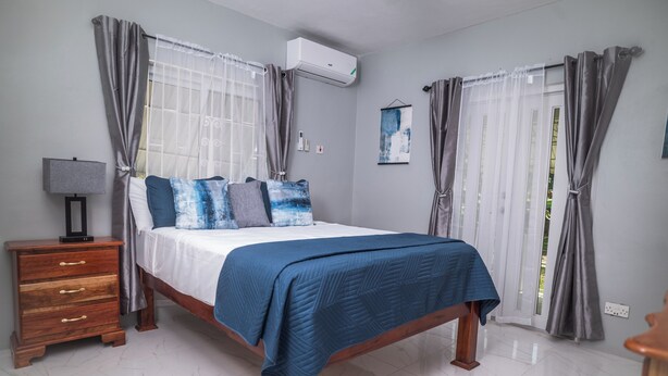 A bedroom with a bed, a nightstand, a lamp, and curtains. - RVR Rentals (Montego Bay)