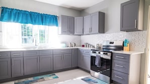 A modern kitchen with grey cabinets, a stainless steel oven, and a window with a blue valance. - RVR Rentals (Montego Bay)