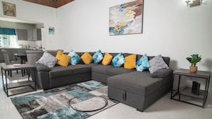 A modern living room with a grey sectional sofa, a patterned rug, and a small side table with a plant. - RVR Rentals (Montego Bay)