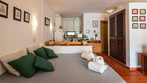 Une chambre d’hôtel avec un lit, des oreillers verts, un lit bien fait avec des serviettes, un coin cuisine avec des placards et un four à micro-ondes, et une porte en bois avec des volets.