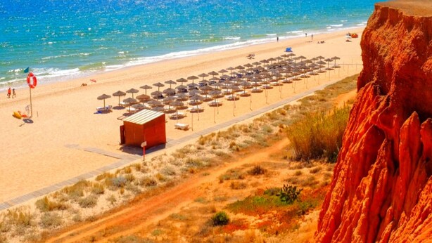 Una playa con un acantilado rojo, una torre de salvavidas y sombrillas.