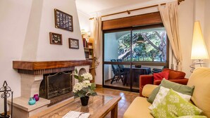 Una sala de estar acogedora con chimenea, una mesa de centro con libros, un sofá con cojines y vistas a un balcón con mobiliario exterior.