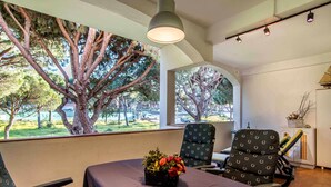 Una habitación con una mesa dispuesta para dos personas, una silla, una mesita con un arreglo floral y vistas a árboles y un estacionamiento a través de amplias ventanas.