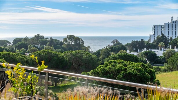 A balcony with a view of the sea, surrounded by greenery and a modern building in the background. - Balaia Residence 2 Bedroom Penthouse With Pool (Albufeira)