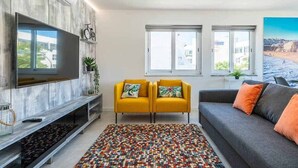 Een woonkamer met een flatscreen televisie, twee gele fauteuils, een grijze bank en een kleurrijk vloerkleed.