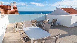 A rooftop terrace with a table and chairs, a view of the sea, and a red-tiled roof.