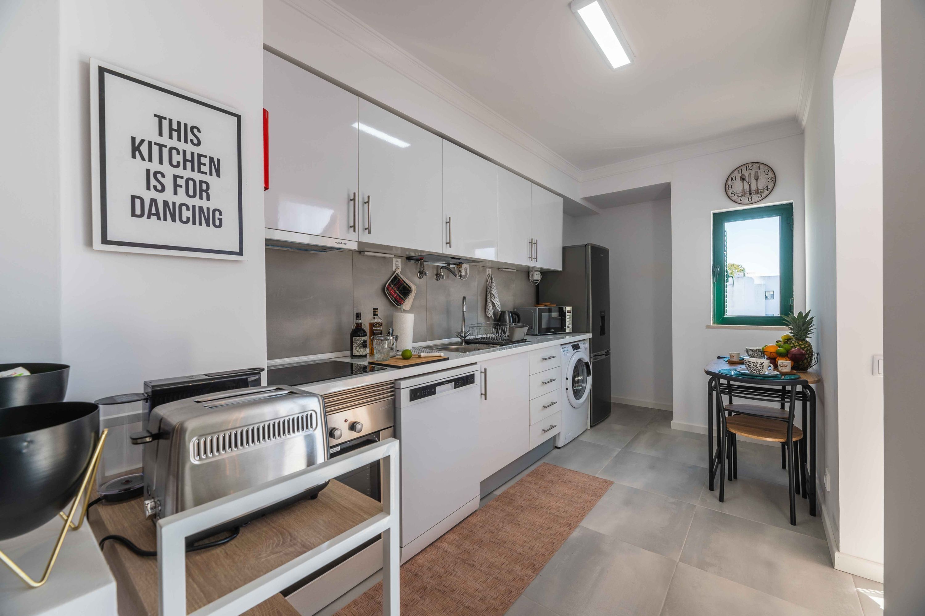 Cocina moderna con armarios blancos, un tostador, un microondas y un lavabo. Hay un reloj en la pared y un cartel enmarcado que dice 'THIS KITCHEN IS FOR DANCING'.