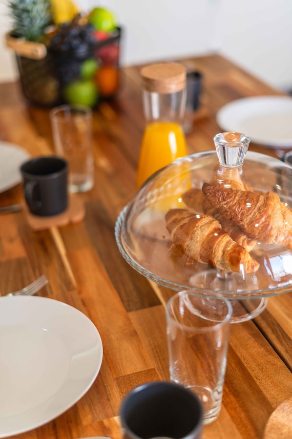 A breakfast setting with croissants on a glass stand, a fruit basket, and a glass of orange juice on a wooden table.