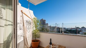 A balcony with a potted plant, a bottle of wine, and two glasses on a wooden table, overlooking a cityscape. - Montechoro Sea View 3 Bedroom Apartment (Albufeira)