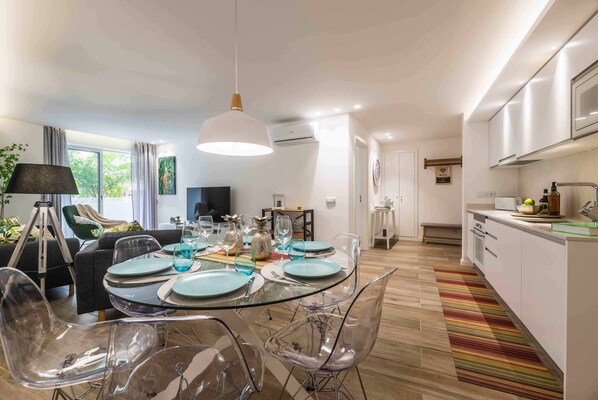 A modern kitchen with white cabinets, a dining area with a glass table and clear chairs, and a large window with curtains.