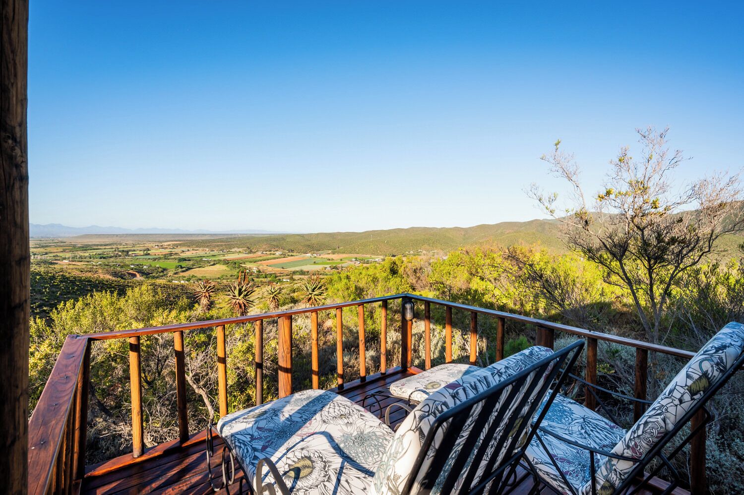 A wooden deck with a railing, a patterned cushion, and a view of a rural landscape with fields and hills.