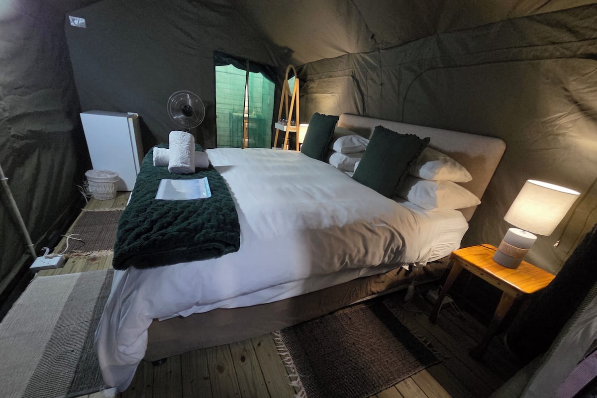 A tent-style room with a bed, bedside table, lamp, and a small table with a fan.