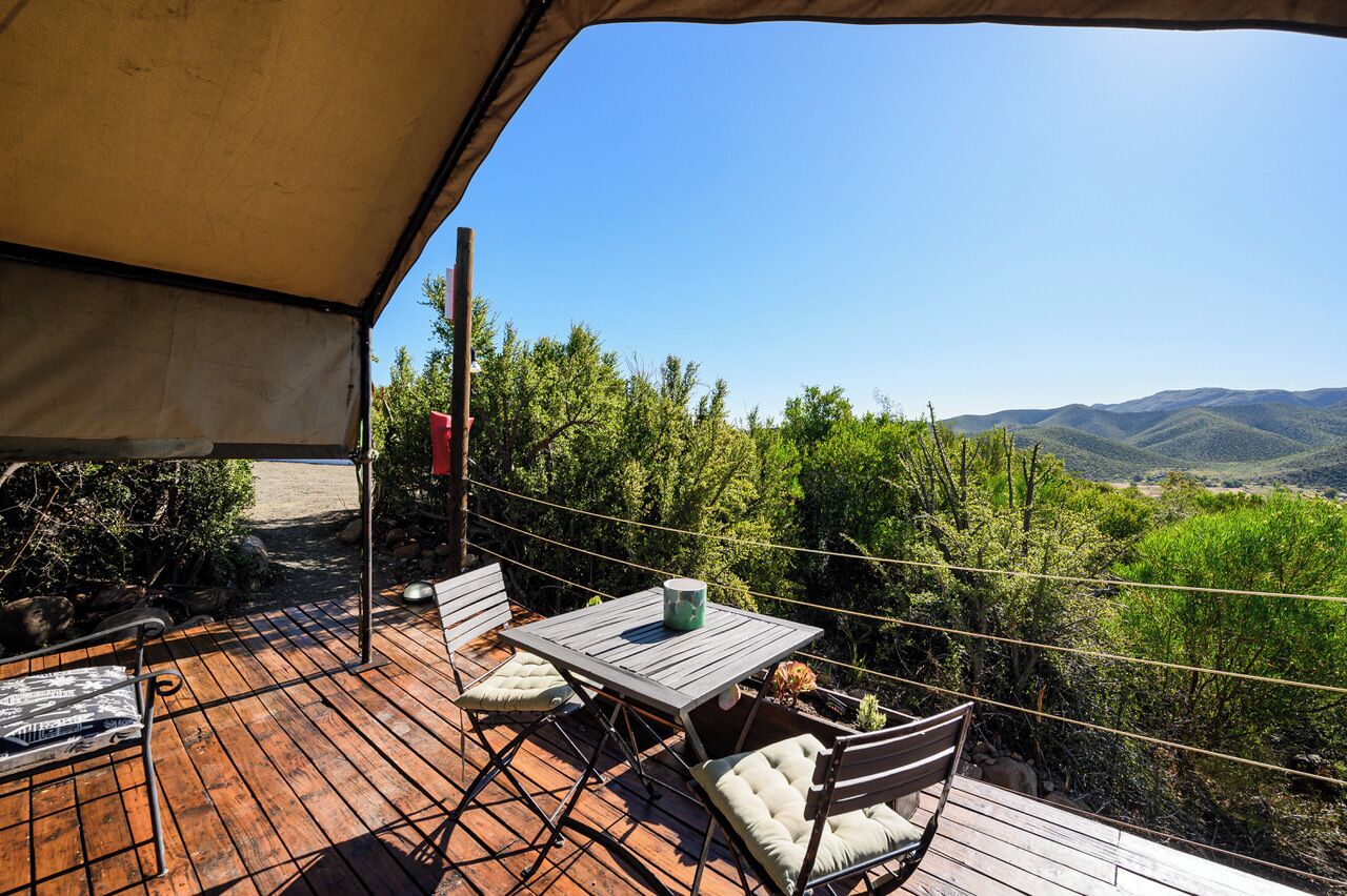 Een buitenterras met een tafel en stoelen, een hanglamp en uitzicht op groene heuvels en bergen.