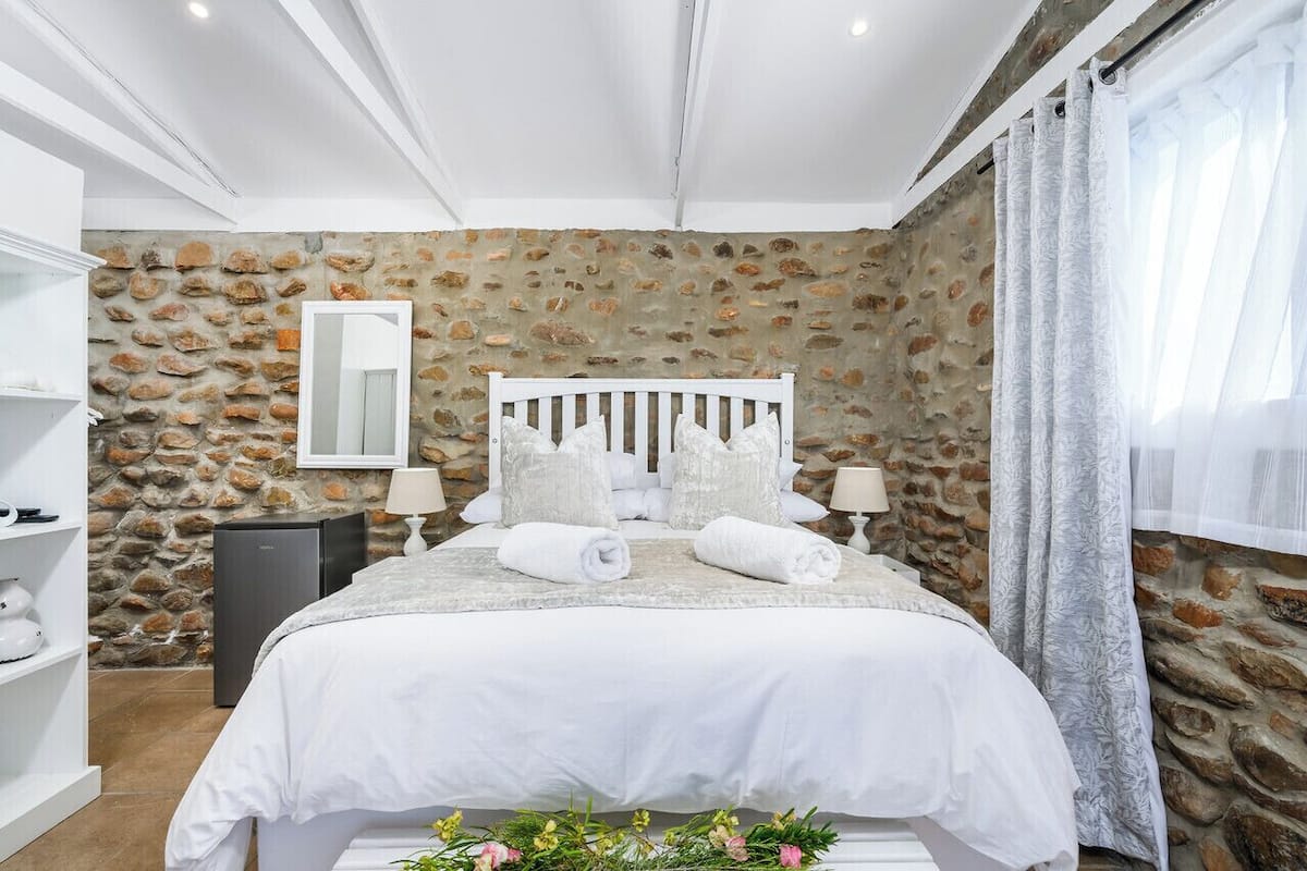 A bedroom with a stone wall, a white bed with pillows, a bedside table, and a mirror.