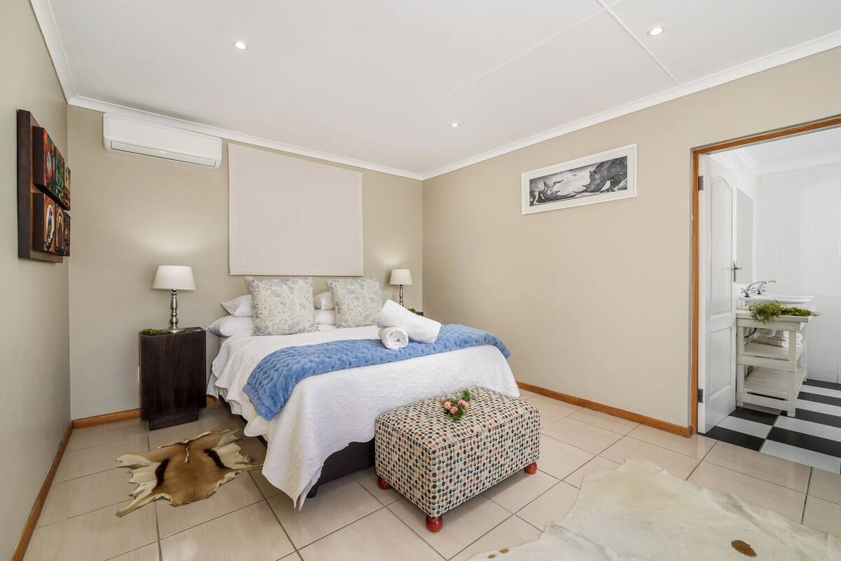 A bedroom with a bed, bedside tables, a rug, a patterned ottoman, and a door leading to a bathroom.