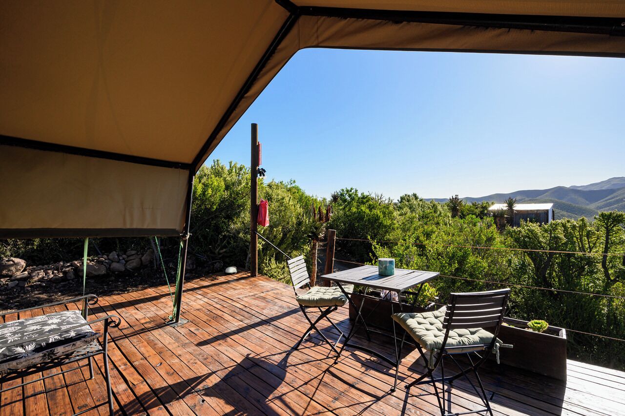 A wooden deck with a table and chairs, a hanging red flag, and a view of green hills and mountains.