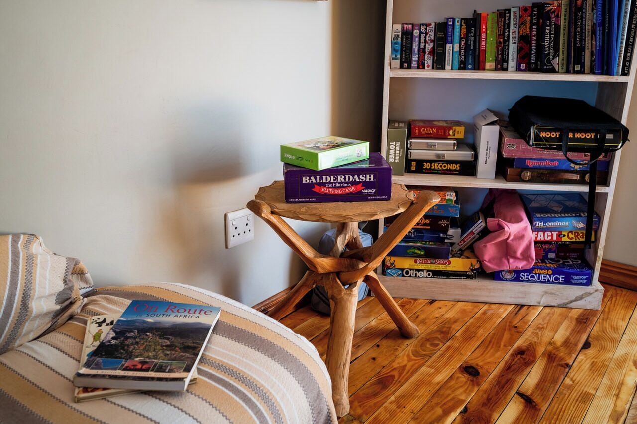 A cozy room with a wooden floor, a striped sofa, a small wooden table with a board game, a bookshelf filled with books and games, and a blanket on the sofa.