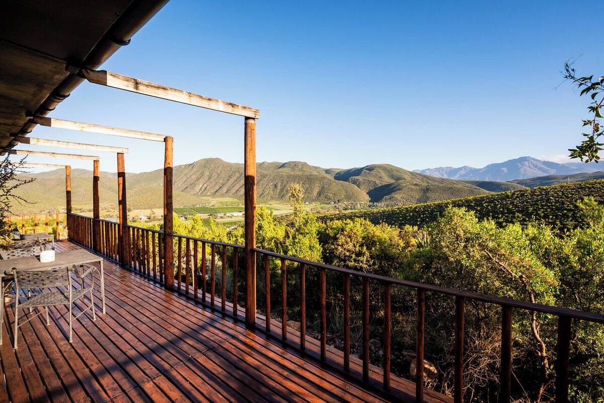 A wooden deck with a table and chairs, overlooking a vineyard and mountains.