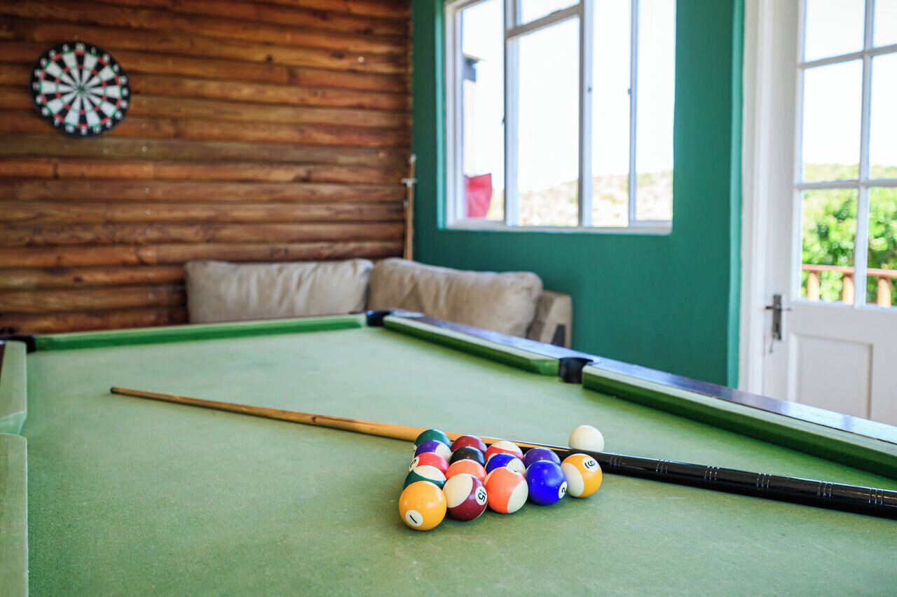 A pool table with balls and a cue stick, a dartboard, and a window with a view of greenery.