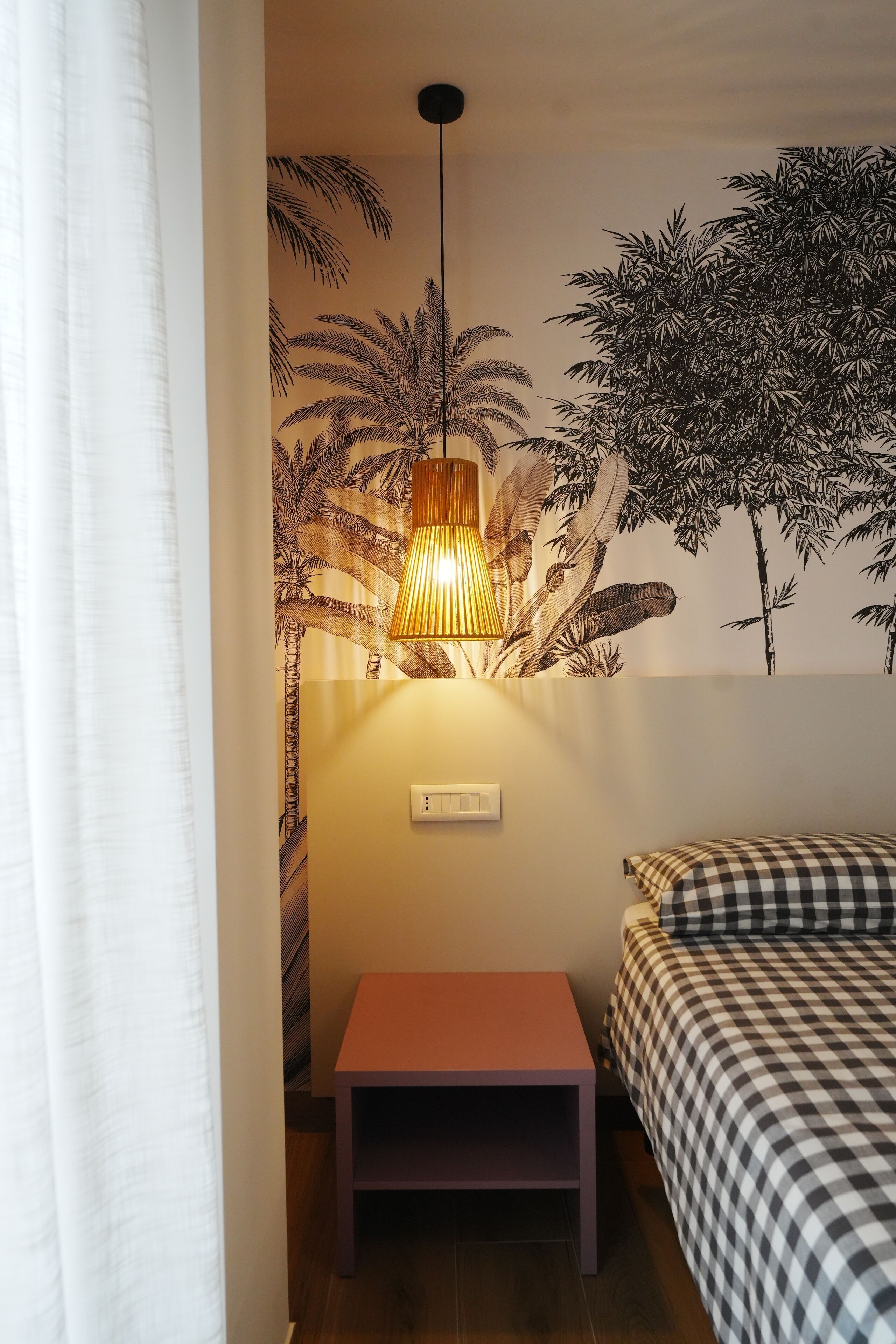 Una habitación con cama, una mesita, una lámpara de pared y un mural de follaje tropical.