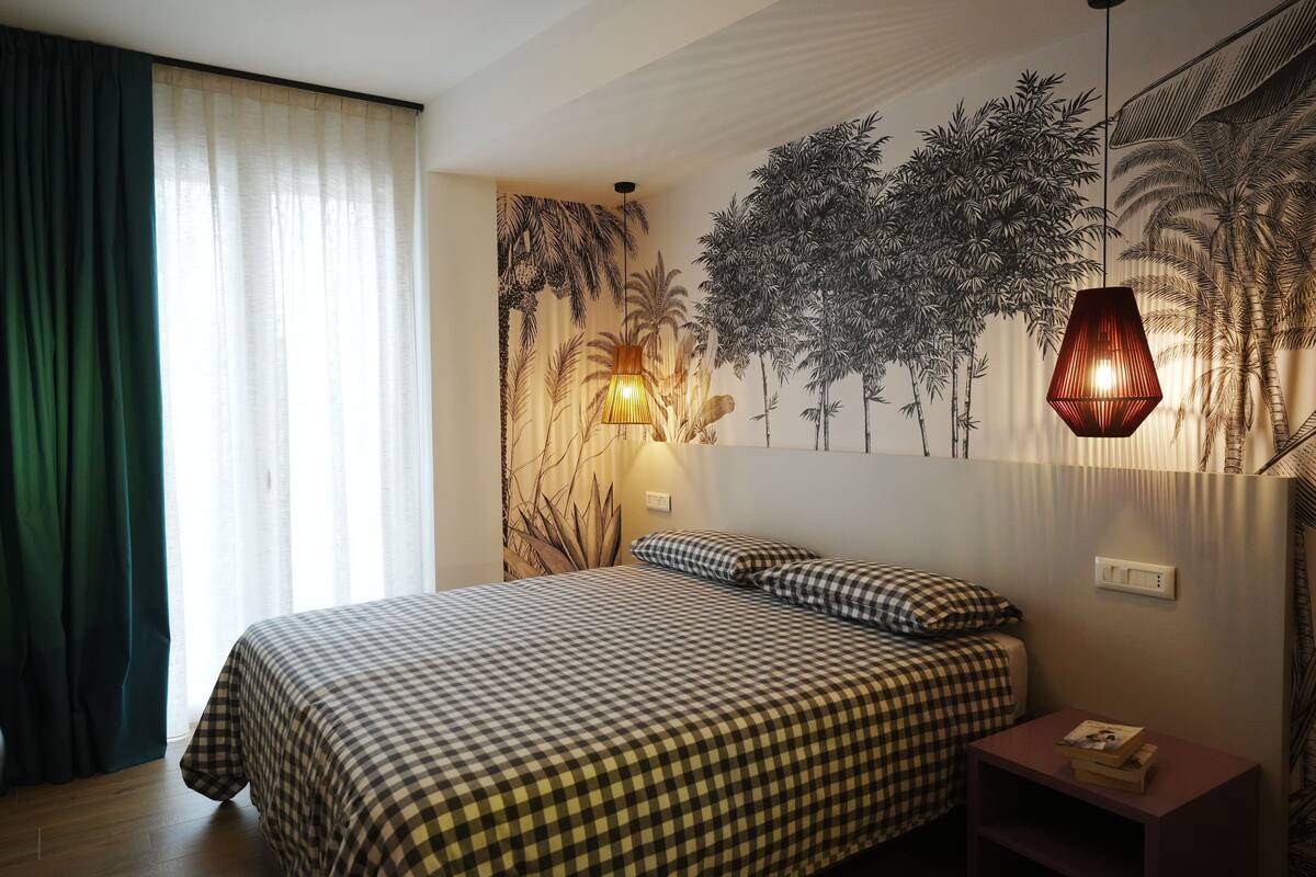 A bedroom with a checkered bedspread, a nightstand with a lamp, and a wall mural of trees.
