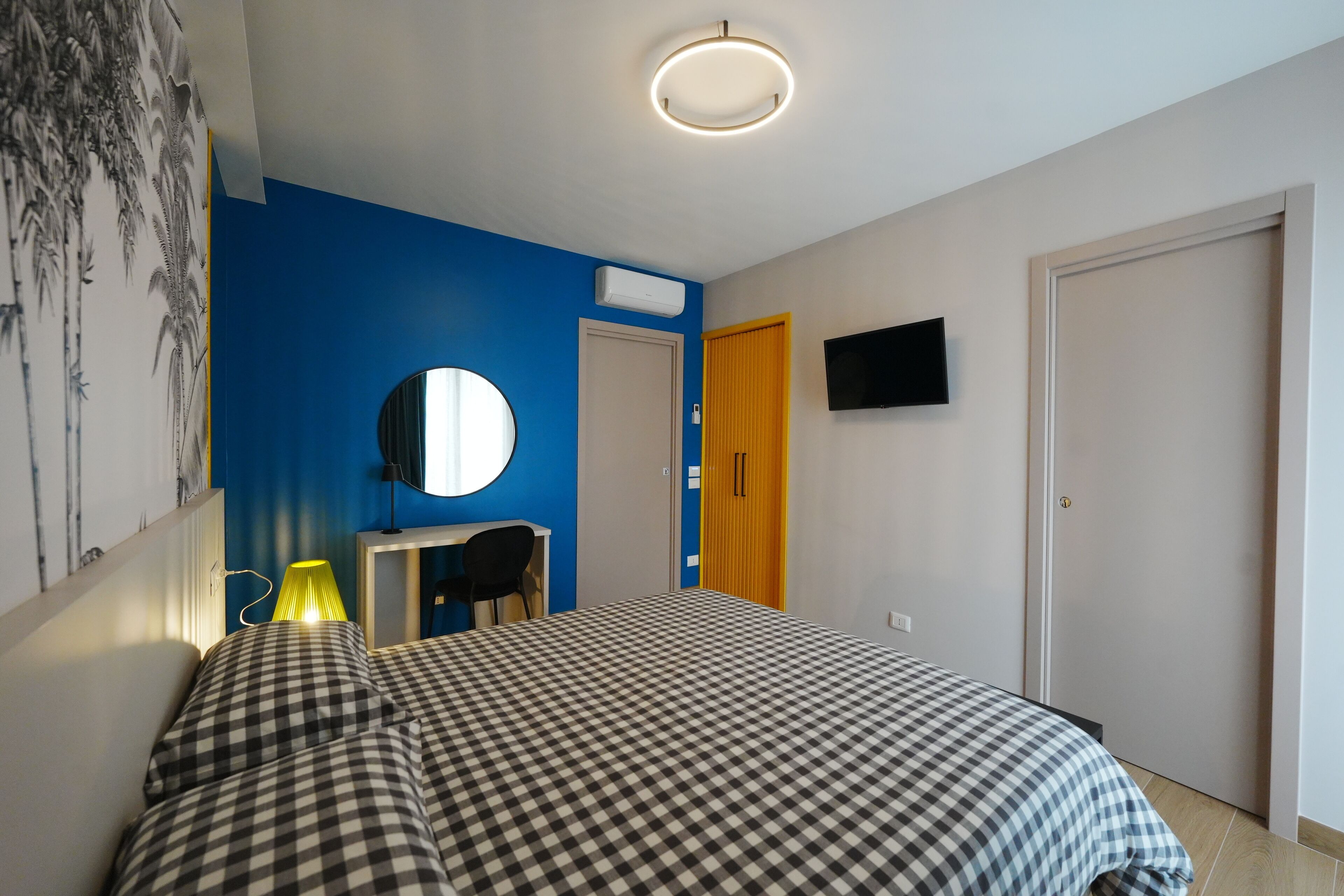 Un dormitorio con colcha a cuadros, un escritorio con lámpara, un televisor montado en la pared y una puerta con panel de madera.
