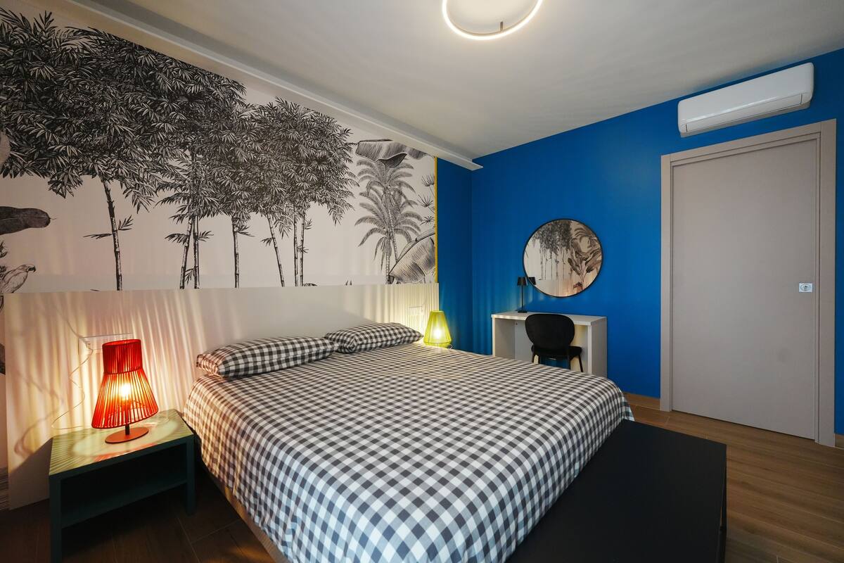 A bedroom with a bed, a nightstand with a lamp, a desk with a chair, a wall mural, and an air conditioning unit.