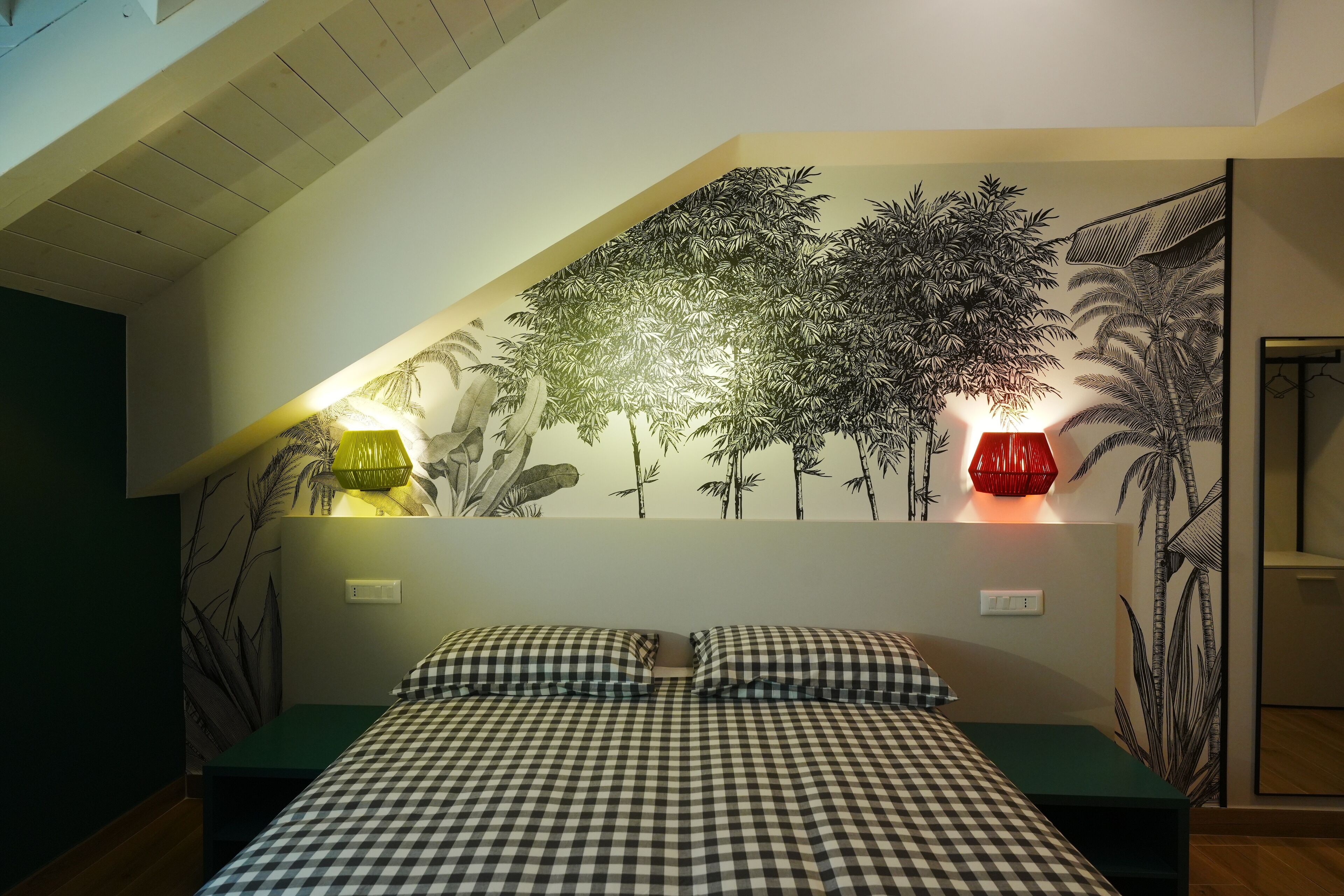 Une chambre avec une couvre-lit à carreaux, deux lampes de chevet et une fresque murale représentant des arbres.