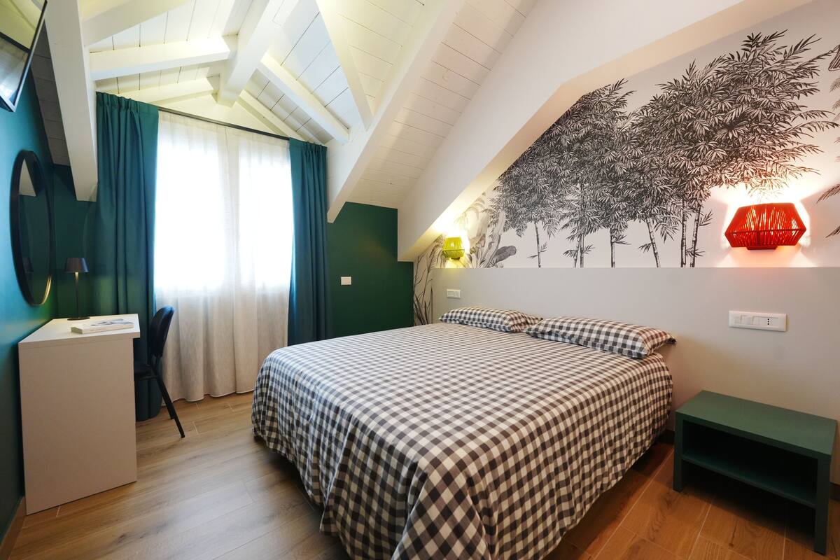 A bedroom with a checkered bedspread, a desk, a chair, a wall-mounted lamp, and a wall mural.