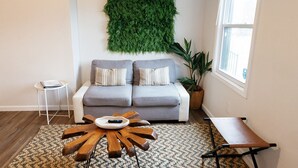 A living room with a grey sofa, two striped pillows, a wooden coffee table, a woven rug, a small white side table, a large green wall hanging, a tall potted plant, and a window with blinds.
