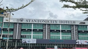 Fachada del Hotel Stanton City con varios comercios, incluyendo One Stop Superstore y un restaurante visible.
