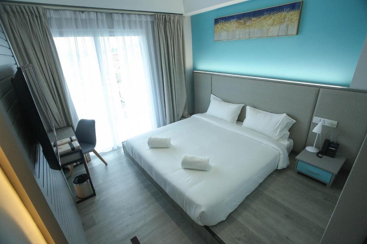 A hotel room with a large bed, a desk, a chair, a nightstand with a lamp, and a painting on the wall.