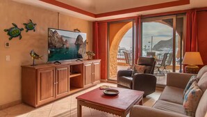 Ein Hotelzimmer mit großem Fernseher, Holzmöbeln, einem Balkon mit Blick auf Meer und Berge sowie einem Deckenventilator.