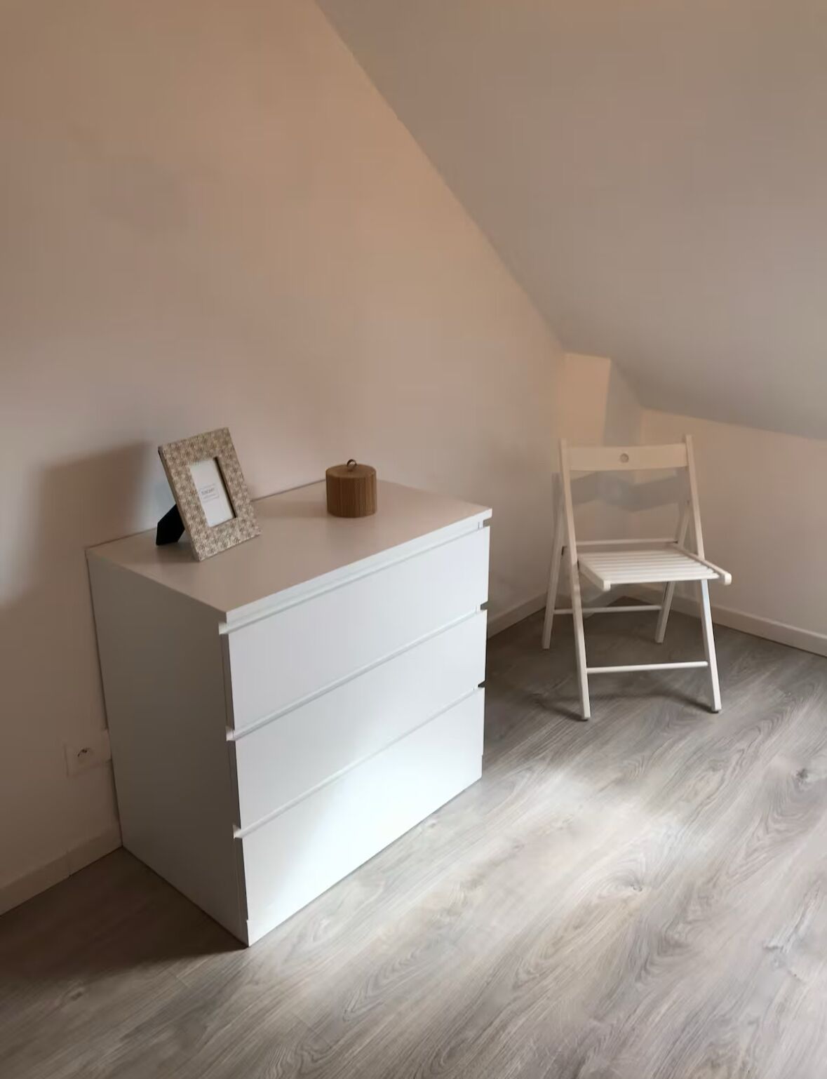 Ein weißer Kommode mit Schubladen, ein kleiner Bilderrahmen und ein zylinderförmiges Objekt darauf. Ein weißer Stuhl steht an der Wand.