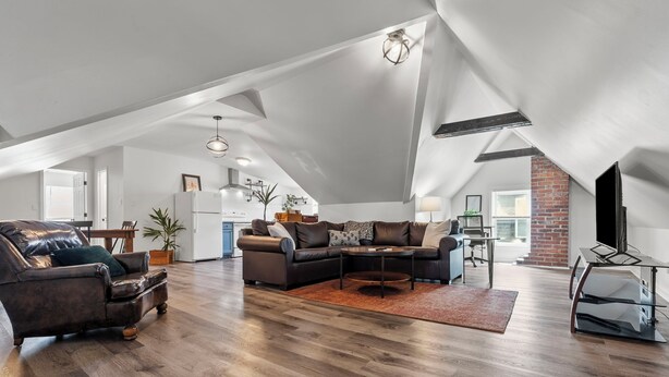 A spacious loft with high ceilings, a large living area with a dark leather sofa, a coffee table, and a dining area with a brick wall and large windows.