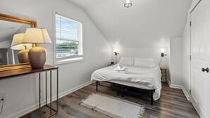 A bedroom with a sloped ceiling, a bed with white bedding, a bedside table with a lamp, and a window with blinds.