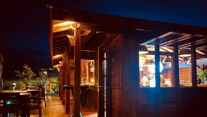 An outdoor wooden deck with tables and chairs, a covered seating area, and a view of the kitchen through a window. - Ecolodge Anacardier- Bungalow Deluxe 1 (Saint-Francois)