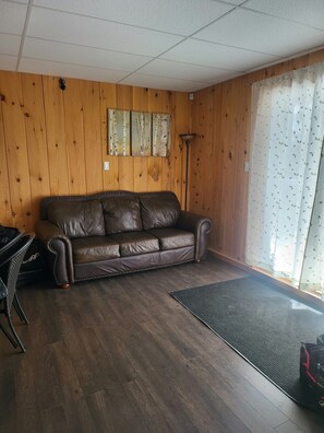 Une pièce avec un canapé en cuir marron, des murs en bois, un lampadaire et une fenêtre ornée d’un rideau à motifs.