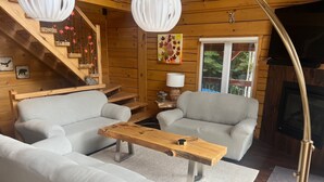 A cozy cabin interior with a sofa set, a wooden coffee table, a fireplace, and a staircase with decorative elements.