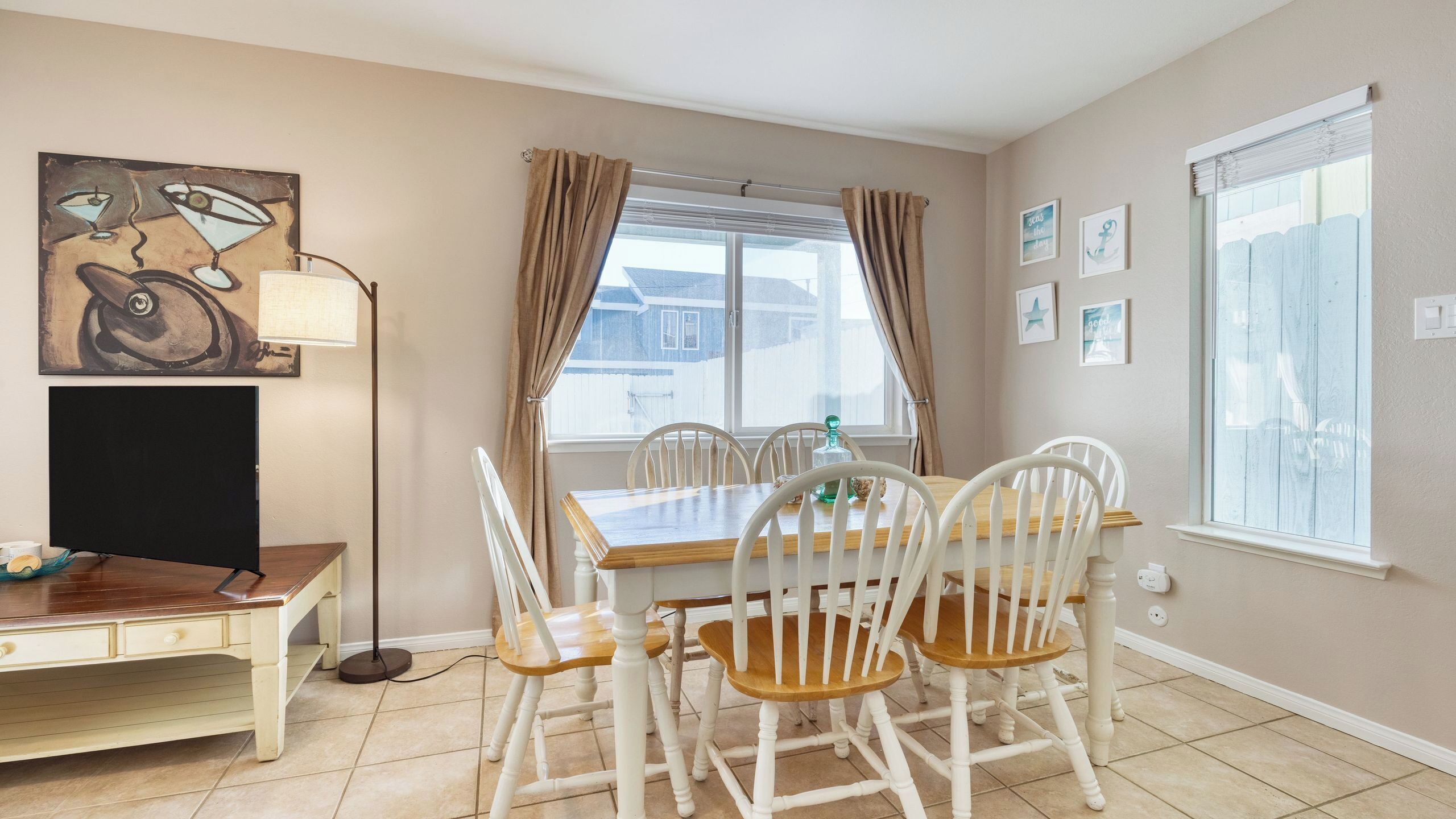 A dining area with a wooden table and white chairs, a painting on the wall, and a window with curtains.