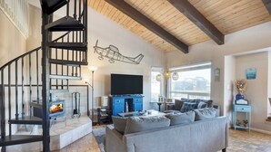 A modern living room with a fireplace, a staircase, a flat-screen TV, and a large window. - Steps to Beach w/ Patio+deck Foosball Happy Bottom Beach House by Avantstay (Oceano)