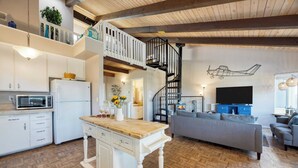 A spacious living area with a kitchen, a wooden floor, a grey sofa, a TV, and a staircase leading to an upper level. - Steps to Beach w/ Patio+deck Foosball Happy Bottom Beach House by Avantstay (Oceano)