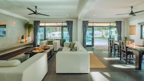 A spacious living area with a sofa, dining table, and chairs, large windows with curtains, and ceiling fans. - Eden Villa A Tropical Paradise Pool Villa (Phuket)
