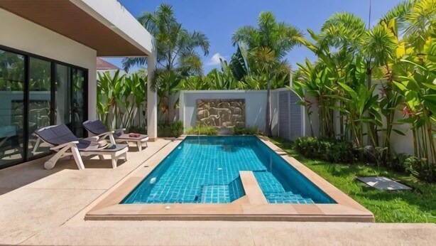 Una piscina rectangular con un muro de piedra, rodeada de un césped cuidado y palmeras.