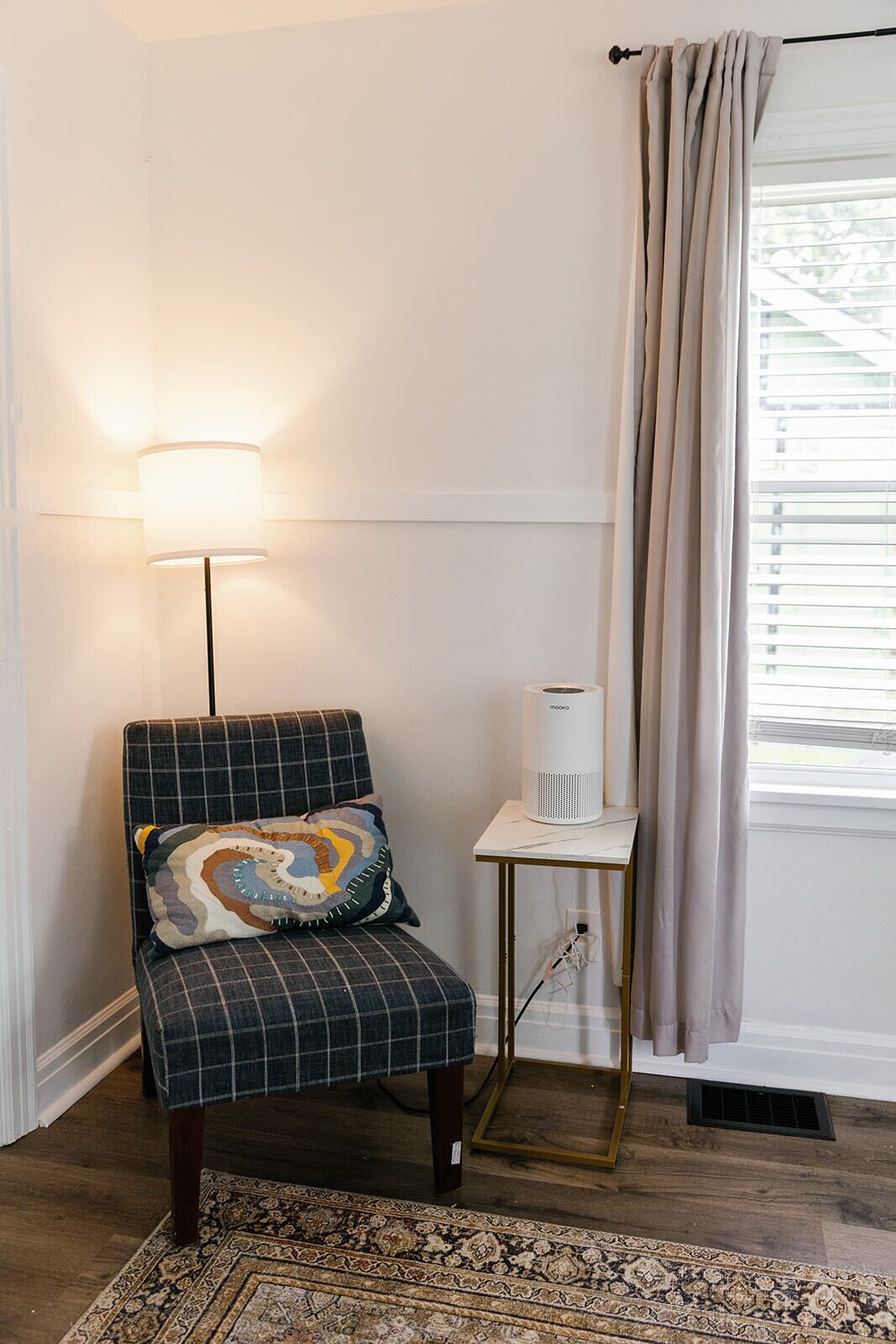 A room with a plaid armchair, a patterned throw pillow, a standing lamp, a small table with a white device, and a window with curtains.